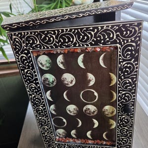 May include: A dark wooden cabinet with a hinged door featuring a moon phase graphic print. The frame is decorated with carved white scrollwork patterns and inlaid with small red stones along the top and bottom edges of the central panel.