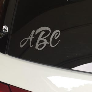 May include: A white car window with a silver decal of the letters "ABC" in a cursive font. The decal is centered on the dark tinted window, with the car's white body and red tail light visible.