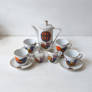 May include: A white ceramic tea set with a whimsical hot air balloon and teddy bear design. The set includes a teapot, sugar bowl, creamer, saucers, and cups. The rims of the cups and saucers are trimmed with gold.