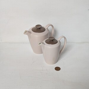 May include: Two ceramic coffee pots with brown lids. The larger pot is light pink and the smaller pot is a slightly darker shade of pink.