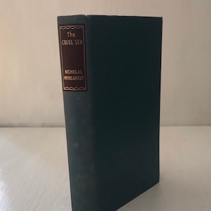 Puede incluir: Un libro de tapa dura titulado "The Cruel Sea" de Nicholas Monsarrat. El libro tiene una cubierta verde oscuro con una etiqueta rectangular en un tono más oscuro de marrón con letras doradas. El libro está de pie sobre una superficie blanca.