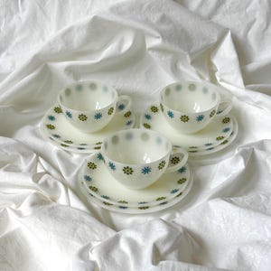 May include: Three white milk glass teacups and matching saucers, each decorated with a repeating pattern of small, stylized star-shaped flowers in alternating shades of olive green and bright blue, arranged in a circular formation.