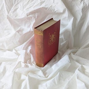 May include: A vintage book with a red cover and gold lettering. The book's spine displays the title "Oliver Twist" in gold script. The cover features an ornate gold design. The pages have a golden hue, suggesting age. The book rests on a white, textured surface.