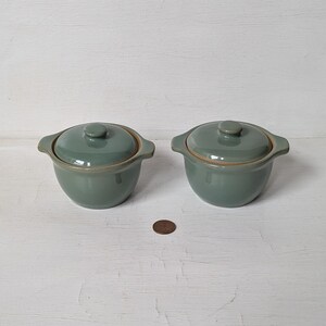 May include: Two small, green ceramic ramekins with lids. The ramekins are round and have a slightly flared lip. The lids are domed and have a small knob on top.
