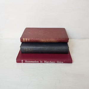 May include: Three vintage books stacked on top of each other. The top book is a dark red with gold lettering and a decorative embossed design. The middle book is black, and the bottom book is a deep red with the title "Automation in Merchant Ships".