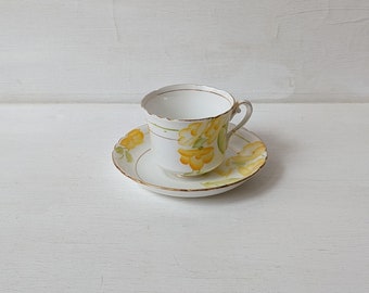 May include: A white teacup and saucer with a yellow floral design. The cup has a gold rim and the saucer has a gold trim.