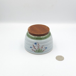 May include: A white ceramic jar with a green and brown lid. The jar features a floral design with blue flowers and red berries.