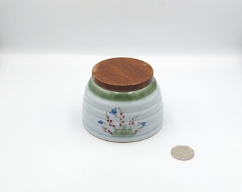 May include: A white ceramic jar with a green and brown lid. The jar has a floral design with blue flowers and red berries.