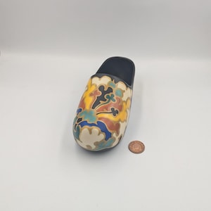 May include: A ceramic shoe shaped like a clog with a black base and a white body. The shoe is decorated with a colorful floral pattern in shades of yellow, blue, green, red, and black.