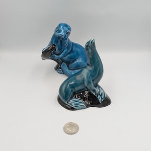 May include: Two blue ceramic sea lion figurines. One sea lion is holding a fish in its mouth. The figurines are sitting on a white surface.