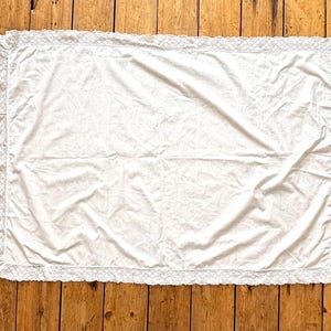 May include: A white rectangular blanket with a delicate lace trim around the edges. The blanket is made of a soft, possibly cotton, material and is slightly wrinkled. The background is a wooden surface.