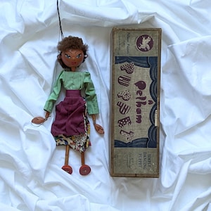 May include: A vintage Pelham puppet doll with a green top and a maroon skirt with a patterned hem. The doll has brown hair and is next to a box with the words "Pelham Puppets" and illustrations of puppets.