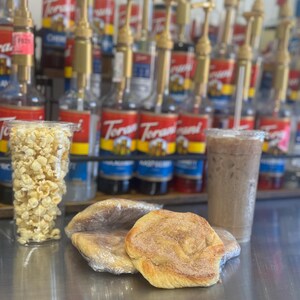 May include: A close-up of a counter with two cinnamon rolls wrapped in plastic, a glass of iced coffee, and a clear cup filled with popcorn. The counter is lined with rows of glass bottles with pumps, some of which have red labels with the word "Torani" printed on them.