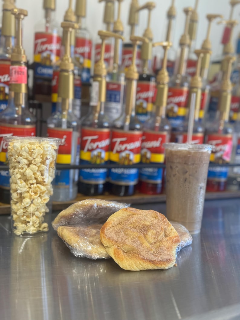 May include: A close-up of a counter with two cinnamon rolls wrapped in plastic, a glass of iced coffee, and a clear cup filled with popcorn. The counter is lined with rows of glass bottles with pumps, some of which have red labels with the word "Torani" printed on them.