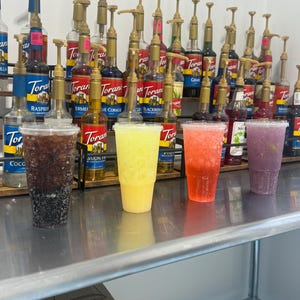 May include: Four different flavored iced drinks in clear plastic cups. The drinks are on a stainless steel counter in front of a shelf with rows of Torani flavored syrups in glass bottles with pumps.