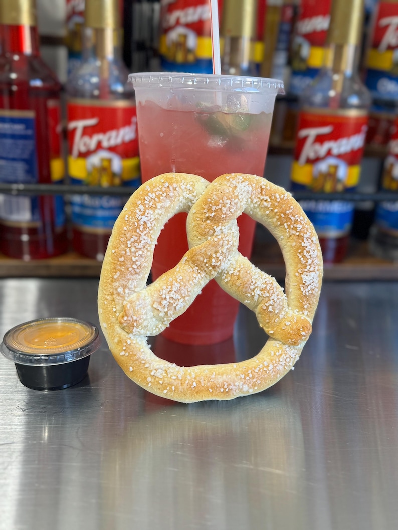May include: A soft pretzel with salt sprinkled on top, a small container of mustard, and a glass of pink lemonade with a straw.