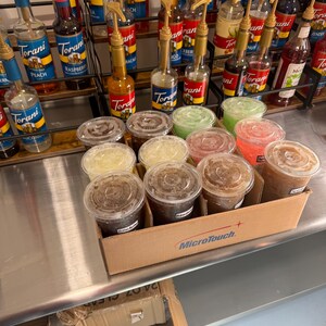 May include: A close-up of a cardboard box filled with clear plastic cups filled with different colored liquids. The box has the words "MicroTouch" printed on it. The cups are arranged in rows and are sitting on a stainless steel surface. There are several bottles of Torani syrups in the background.