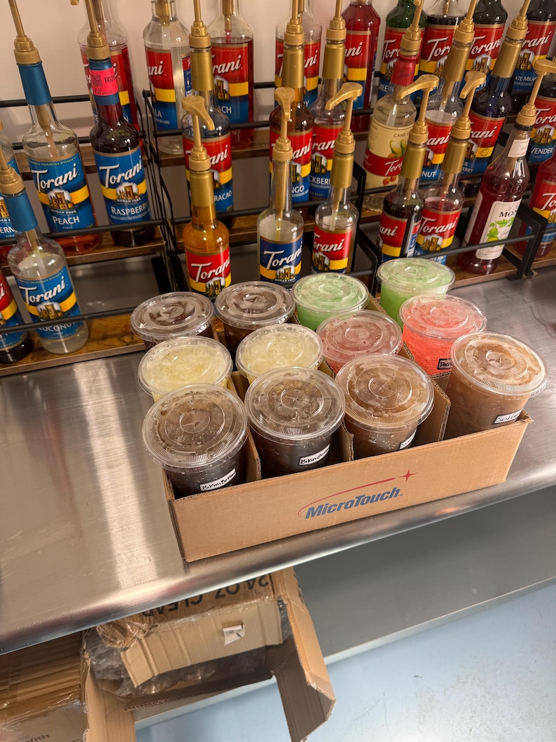 May include: A close-up of a cardboard box filled with clear plastic cups filled with different colored liquids. The box has the words "MicroTouch" printed on it. The cups are arranged in rows and are sitting on a stainless steel surface. There are several bottles of Torani syrups in the background.