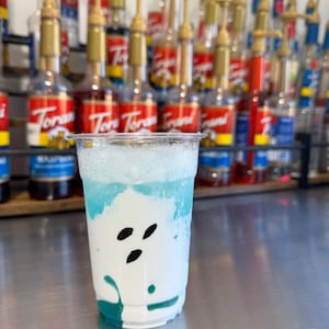 May include: A clear plastic cup with a white and blue layered drink. The drink has a white foam top and a blue base. The cup is sitting on a silver surface. There are rows of bottles with red and gold labels in the background.