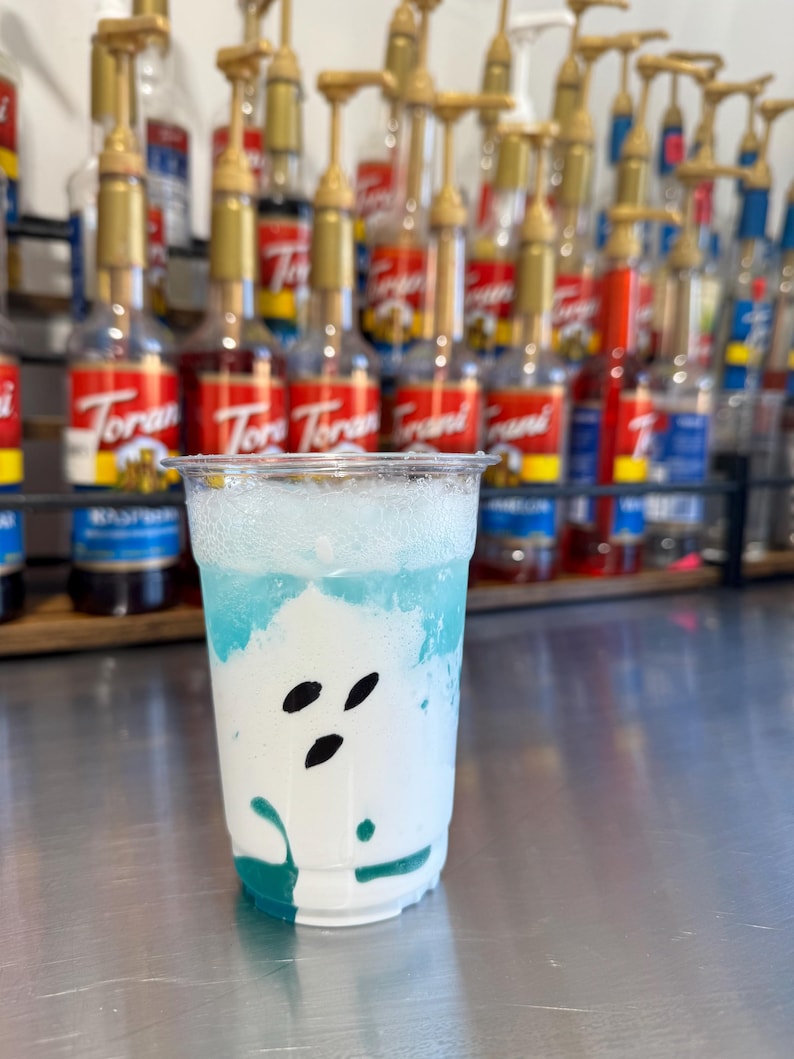 May include: A clear plastic cup with a white and blue layered drink. The drink has a white foam top and a blue base. The cup is sitting on a silver surface. There are rows of bottles with red and gold labels in the background.