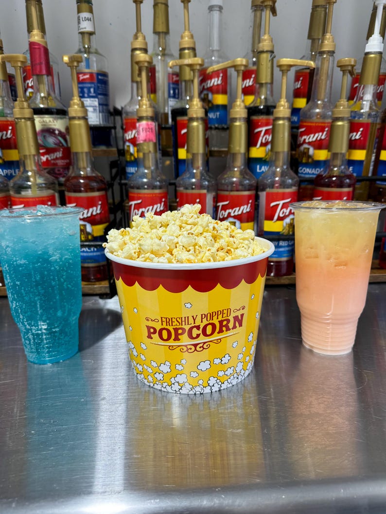 May include: A yellow and red popcorn bucket with the text "FRESHLY POPPED POPCORN" on the front. The bucket is filled with popcorn and is sitting on a silver surface. There are two clear cups with blue and orange liquids next to the bucket.