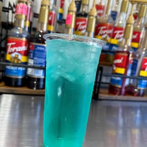 May include: A tall clear glass filled with a bright blue, carbonated beverage with ice. The glass is sitting on a stainless steel surface.