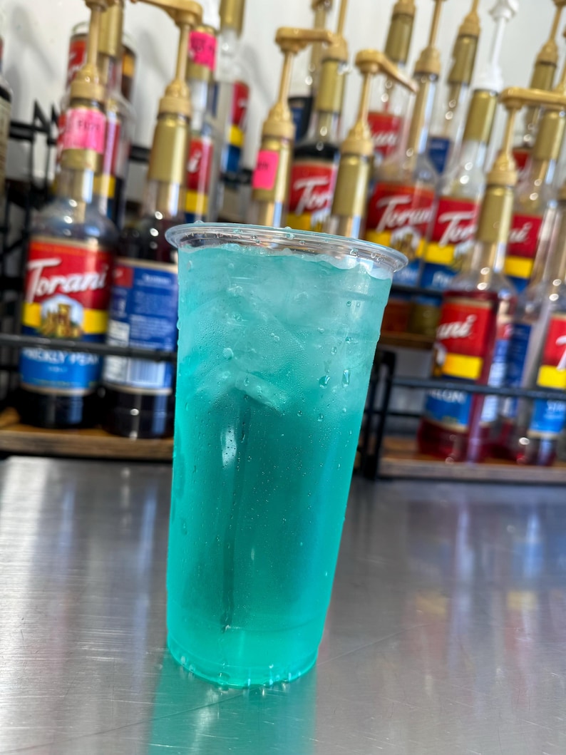 May include: A tall clear glass filled with a bright blue, carbonated beverage with ice. The glass is sitting on a stainless steel surface.