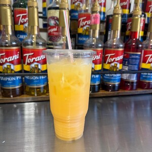 May include: A clear plastic cup filled with orange juice and ice. The cup is sitting on a stainless steel surface. There are several bottles of flavored syrups in the background.