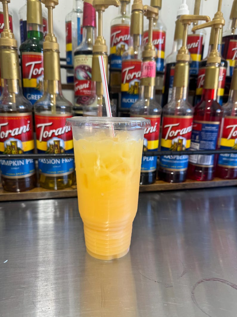 May include: A clear plastic cup filled with orange juice and ice. The cup is sitting on a stainless steel surface. There are several bottles of flavored syrups in the background.