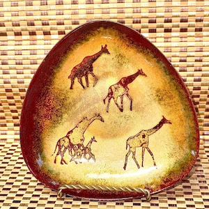 May include: A triangular enameled metal dish featuring a gradient of deep red at the edges transitioning to a golden yellow center. The surface is decorated with five dark brown giraffe silhouettes, including one small giraffe standing near a larger one.