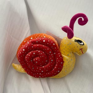 May include: A soft toy snail with a red and orange shell, yellow body, and pink antennae. The shell is decorated with silver glitter spots. The snail has a smiling face with black eyes and a red mouth. A tag is attached to the toy.