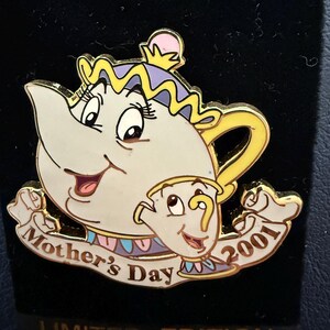 Può includere: Spilla in edizione limitata Mother's Day 2001 con una teiera e una tazza dei cartoni animati. La spilla ha un corpo bianco con dettagli gialli, viola e rosa. Il testo "Mother's Day" è su uno striscione.