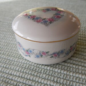Puede incluir: Una caja de porcelana blanca para alhajas con un borde dorado y un dise&ntilde;o floral. La tapa presenta un dise&ntilde;o floral en forma de coraz&oacute;n con flores rosas y azules.