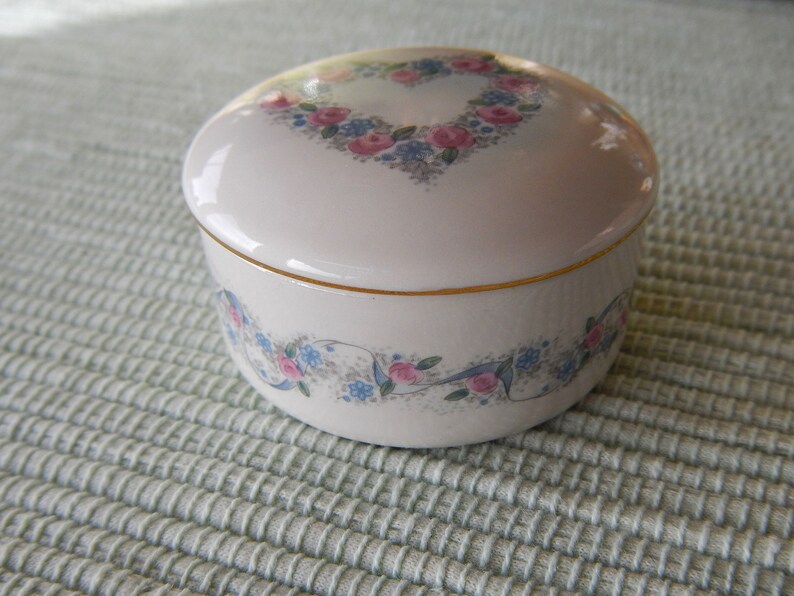 Puede incluir: Una caja de porcelana blanca para alhajas con un borde dorado y un dise&ntilde;o floral. La tapa presenta un dise&ntilde;o floral en forma de coraz&oacute;n con flores rosas y azules.