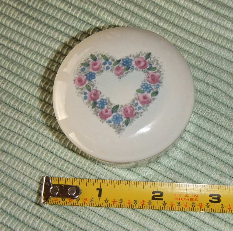 Puede incluir: Caja de cer&aacute;mica blanca para guardar peque&ntilde;os objetos con un dise&ntilde;o de coraz&oacute;n floral. El coraz&oacute;n est&aacute; hecho de flores rosas y azules con hojas verdes. La caja tiene un di&aacute;metro de aproximadamente 7,6 cm.