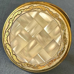 May include: A round, vintage compact with a gold-colored metal frame. The center features a woven pattern of rectangular mother-of-pearl pieces. A decorative gold filigree border encircles the mother-of-pearl.