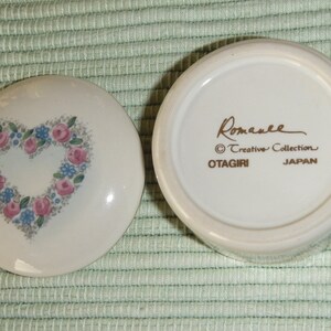 Puede incluir: Caja de cer&aacute;mica blanca para alhajas con un dise&ntilde;o en forma de coraz&oacute;n con flores rosas y azules. La tapa tiene las palabras "Romanee Creative Collection OTAGIRI JAPAN" en letras doradas.