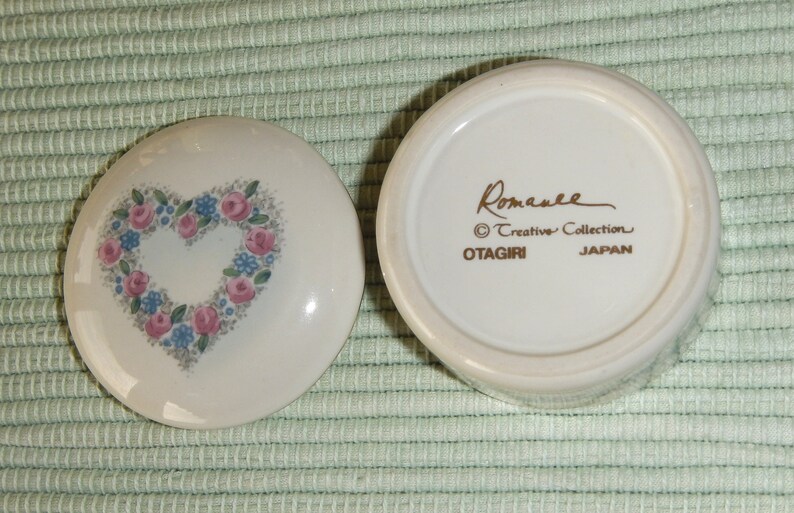Puede incluir: Caja de cer&aacute;mica blanca para alhajas con un dise&ntilde;o en forma de coraz&oacute;n con flores rosas y azules. La tapa tiene las palabras "Romanee Creative Collection OTAGIRI JAPAN" en letras doradas.