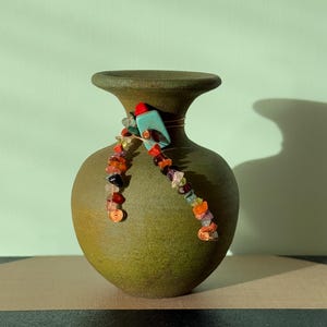May include: A green ceramic vase with a rounded body and a flared rim. A decorative strand of colourful gemstones and copper wire is wrapped around the neck. A turquoise and red accent piece is attached to the wire.