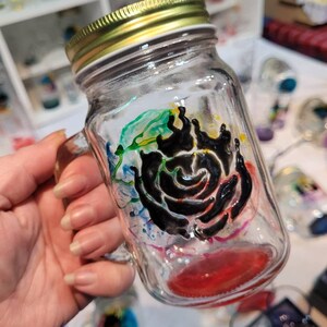 May include: A clear glass mason jar mug with a black and white rose design. The rose is surrounded by colourful paint splatters in shades of blue, green, yellow, and red. The jar has a gold lid and a handle.