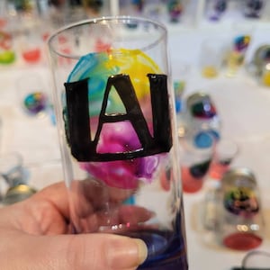 May include: A clear glass shot glass with a colourful abstract design and the letter "A" painted in black on the front.