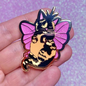 May include: Gold-tone enamel pin featuring a black and orange tabby cat with pink butterfly wings. The cat is wearing a crown and has a paw print on its tail.