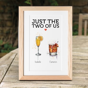 Peut inclure: Une impression encadrée avec le texte "Just the two of us" et un symbole de cœur. Deux cocktails sont illustrés sous le texte, étiquetés "Isabelle" et "Cameron".
