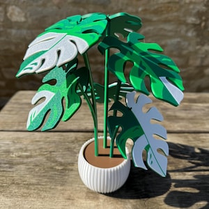 Könnte beinhalten: Eine dekorative künstliche Monstera-Pflanze in einem weißen, gerippten Topf. Die Pflanze hat große, grüne Blätter mit weißen Akzenten. Der Topf steht auf einer Holzoberfläche. Ein lebendiges Wohnaccessoire.