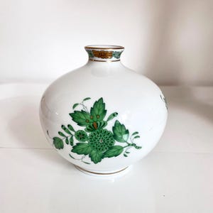 Puede incluir: Un jarrón de cerámica blanca con cuerpo redondo y cuello estrecho. El jarrón está decorado con diseños florales verdes y ribetes dorados alrededor del cuello y la base. El diseño floral presenta hojas verdes y una flor central.