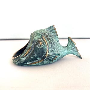 Op de afbeelding: Een groene en gouden metalen asbak in de vorm van een vis. De vis heeft een gedetailleerd ontwerp en ligt op zijn zij.