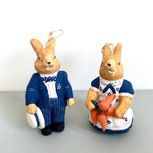 Könnte beinhalten: Zwei Keramik-Kaninchenfiguren. Ein Kaninchen trägt einen blauen Anzug mit Fliege und hält einen Hut. Das andere Kaninchen trägt ein blau-weißes Kleid und eine Schürze und hält Karotten. Beide haben hellbraunes Fell.