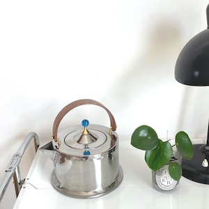 May include: A polished stainless steel kettle with a cylindrical body, a curved wooden handle, and a lid featuring a small blue spherical knob. The kettle has a short spout and a flat base, reflecting light on its metallic surface.