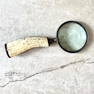 May include: A vintage magnifying glass with a cream-colored handle and a black rim around the glass. The handle has a textured, organic pattern. The magnifying glass is a classic tool for reading and examining small details.