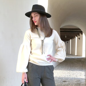 Puede incluir: Una mujer con un suéter blanco con un collar largo de cuentas, pantalones grises y un sombrero fedora negro. Lleva un bolso negro y está de pie en un pasillo blanco con arcos.